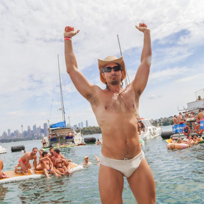 A man in a cowboy hat and white briefs stands with arms raised on a boat, surrounded by people swimming and relaxing on boats and rafts, with a city skyline visible in the background under a partly cloudy sky.