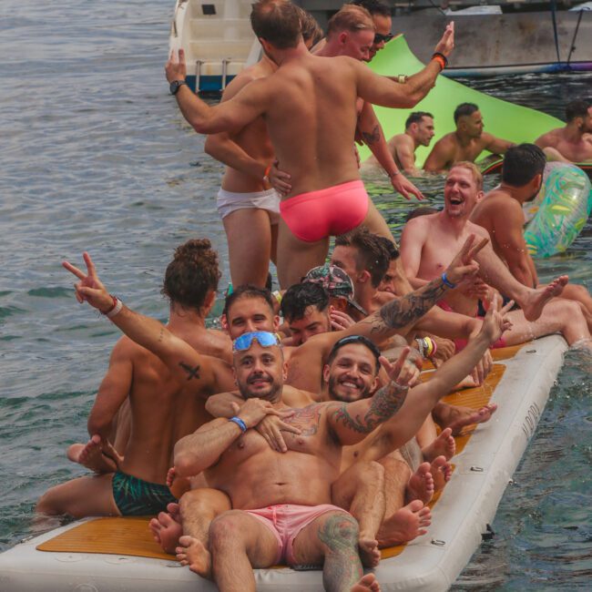 A group of men in swimsuits sit and pose cheerfully on an inflatable dock in the water, some making peace signs, with others swimming or lounging nearby and a boat in the background.