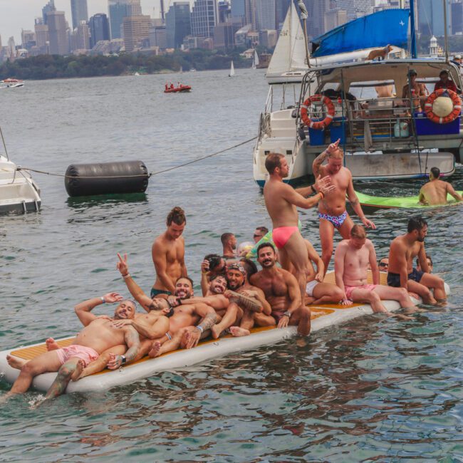 A group of men in swimwear pose and smile on a floating platform in the water, with sailboats and a city skyline in the background. The mood is festive and joyful.