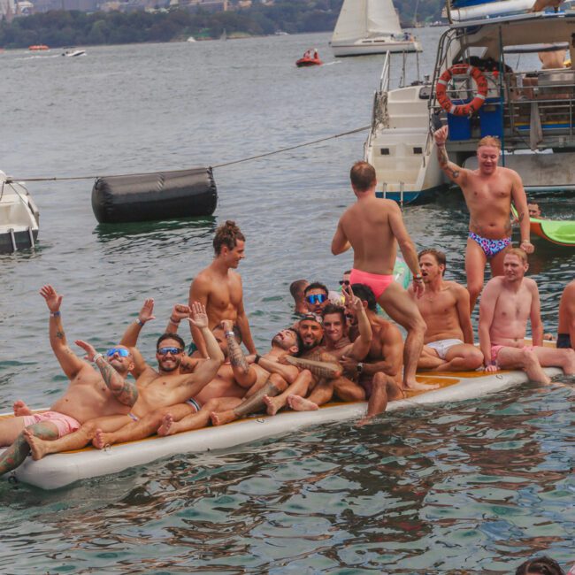A large group of men in colorful swimwear sit and pose on a floating mat in the water, waving and smiling, with boats and a city skyline visible in the background.