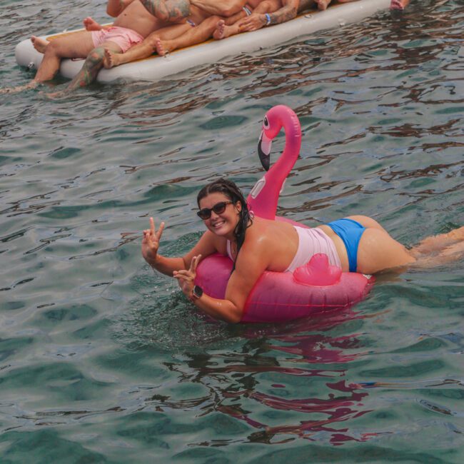 A smiling person in swimwear lounges on a pink flamingo float in the water, flashing peace signs. In the background, a group of people relaxes together on a large float. The atmosphere is festive and joyful.