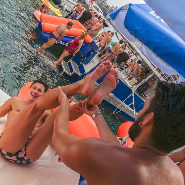 Two people playfully slide on an inflatable float in the water near boats crowded with people. The scene is lively, with bright colors, festive atmosphere, and visible water and sky in the background.