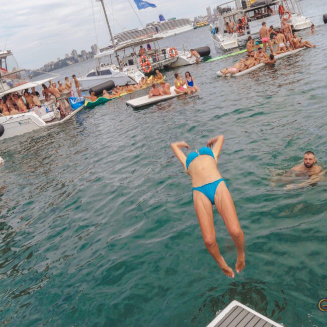 A person in a blue swimsuit dives into the water from a dock, surrounded by people swimming and relaxing on boats in a lively, social scene. The sky is cloudy and the background shows city buildings.