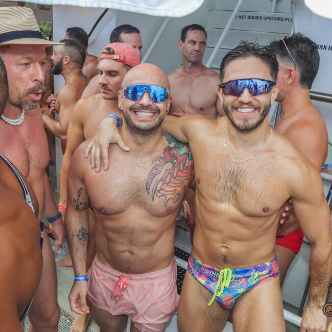 A group of shirtless men wearing swim trunks and sunglasses smile and pose together on a boat deck during a lively pool party. Two men in the center have their arms around each other and are cheerfully facing the camera.