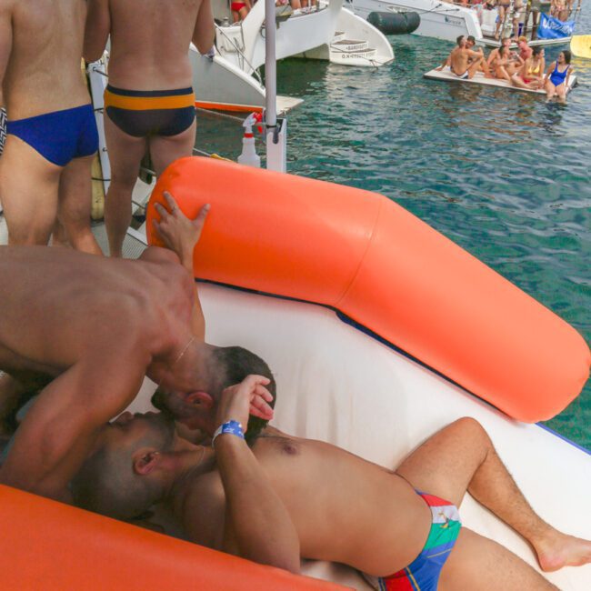 Two men are kissing on an orange inflatable raft on the water, surrounded by other men in swimwear. Boats and more people are visible in the background, suggesting a lively party or event.