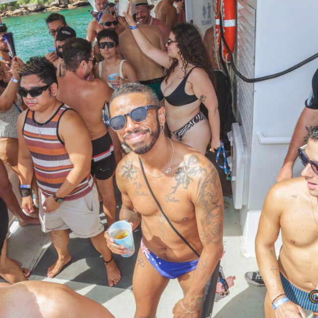 A group of people in swimsuits enjoy a lively party on a boat under sunny weather, some holding drinks and dancing, with blue water and greenery visible in the background.