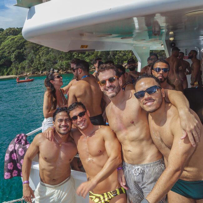A group of four men in swimwear pose and smile on the deck of a boat, surrounded by other people enjoying a sunny day by the water with lush greenery in the background.