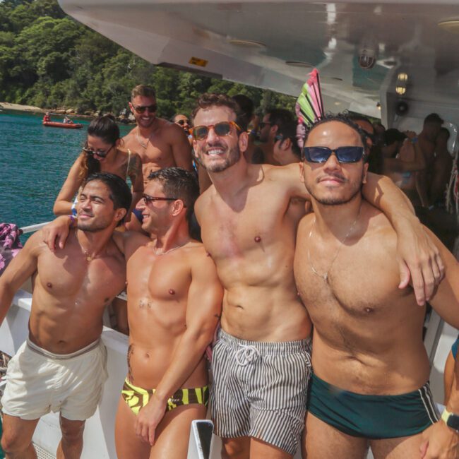 Four men in swimwear pose and smile on a boat deck with other people in the background. The boat is on the water near a forested shoreline, and it is a sunny day. The atmosphere is festive and relaxed.