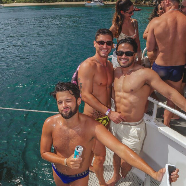 Three men in swimwear stand on a boat’s ladder smiling at the camera, with lush green trees and blue water in the background. One man holds a drink can, and other people are seen relaxing on the boat.