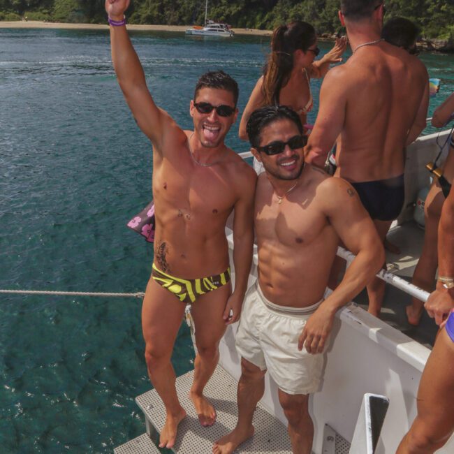 Two smiling men in swimwear stand on boat steps; one raises his arm in celebration. Behind them, others mingle on the boat, with blue water and a forested shore in the background.