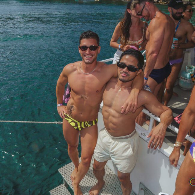 Two men in swimwear smile and pose together on a boat, with one man’s arm around the other. Several other people in swimsuits are behind them, and blue water is visible in the background.