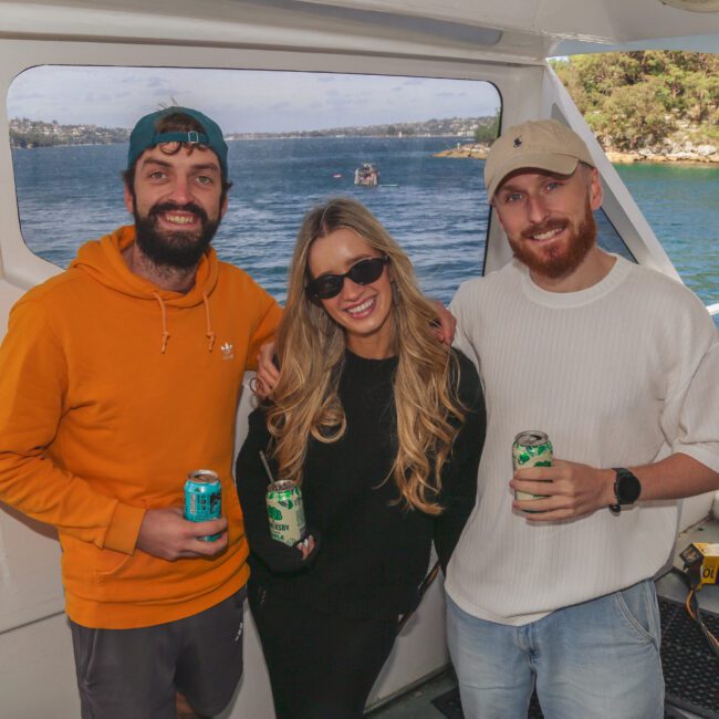Three people stand smiling on a boat with drinks in hand. Water and a forested shoreline are visible in the background. The two men wear casual clothes and hats; the woman, in the center, wears black sunglasses and a black jacket.