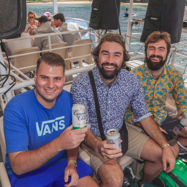 Three smiling men sit on a boat holding cans of drink, with water and a beach visible in the background. Other people relax on seats nearby, and bags are seen around them.