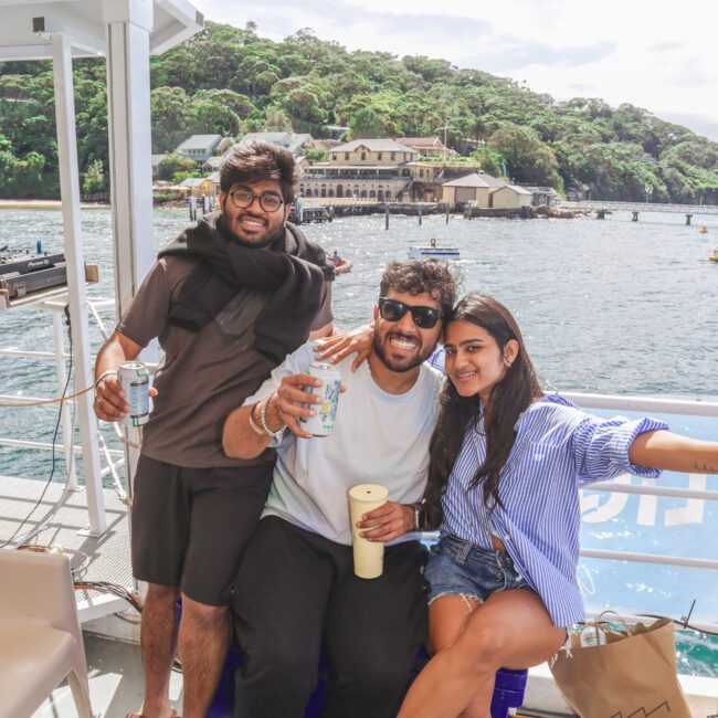 Three friends are smiling and posing together on a boat, holding drinks. There's water and lush green hills with a building in the background under a bright, sunny sky.