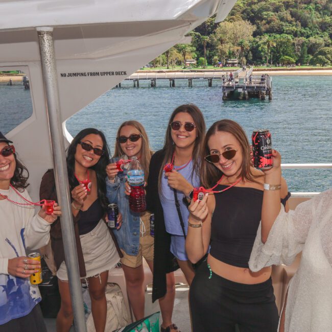 Six women smiling and holding bubble wands on a boat, with water and a lush, green shoreline in the background. They are wearing sunglasses and casual outfits, enjoying a sunny day together.