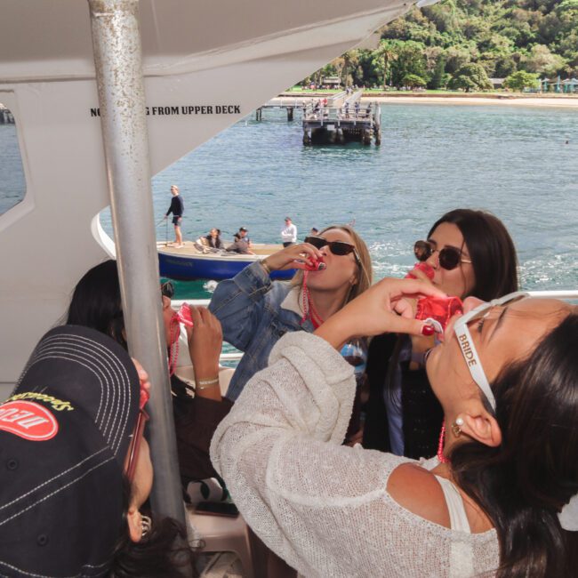 A group of women on a boat cheerfully take shots together, surrounded by water and lush greenery in the background, with a dock and another boat visible nearby.