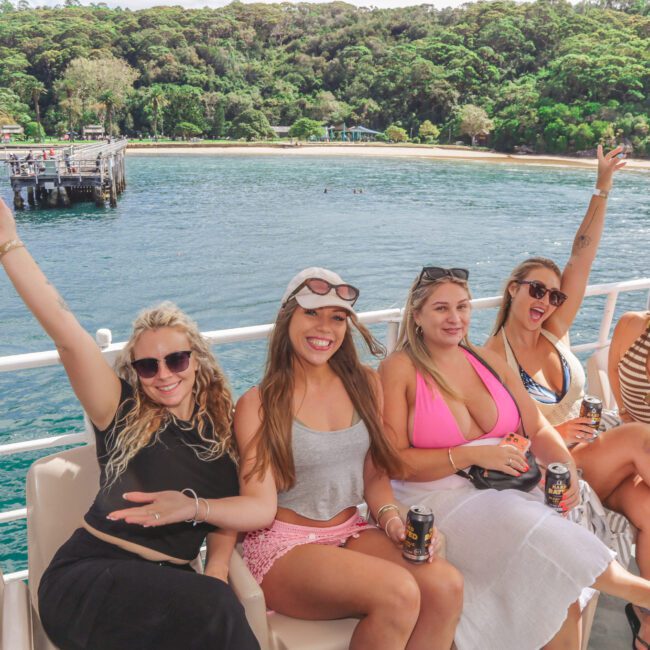 Five women sit on a boat, smiling and waving at the camera, with drinks in hand. Behind them, there is a blue body of water, a pier, and lush green trees along the shoreline under a clear sky.