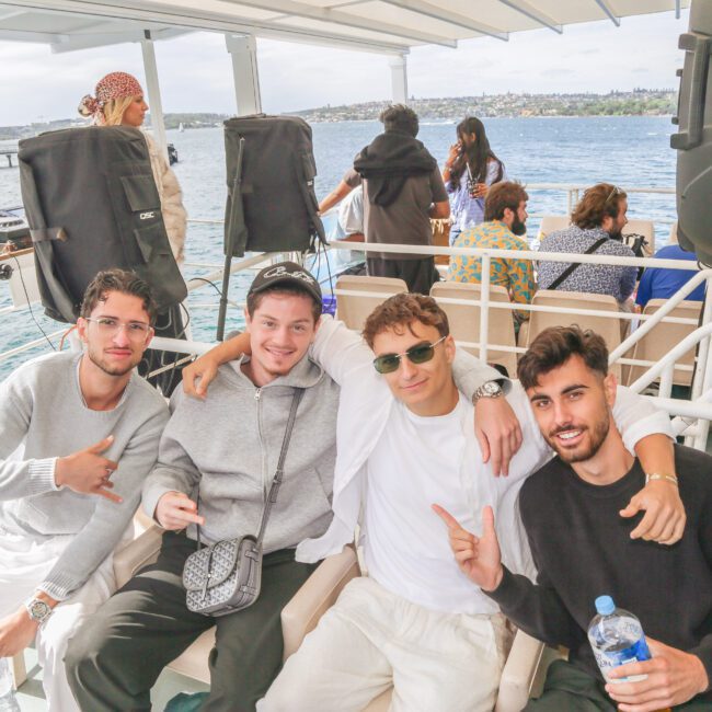 Four young men sit together on a boat, smiling and posing for the camera. The sea and distant shore are visible behind them, and other people are seated in the background. Audio equipment and water bottles are also visible.
