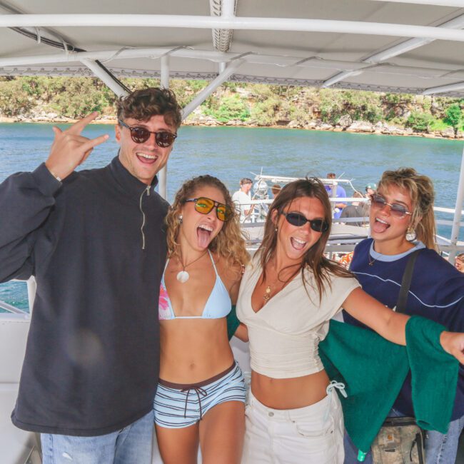 Four young adults pose happily on a boat, smiling and making celebratory gestures. One person holds a bottle of wine, and the background shows water, trees, and people on another boat.