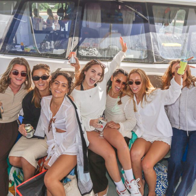 A group of seven women sit together on a boat, smiling and laughing, holding drinks and dressed in casual summer outfits. The atmosphere is festive and joyful, with a boat in the background.
