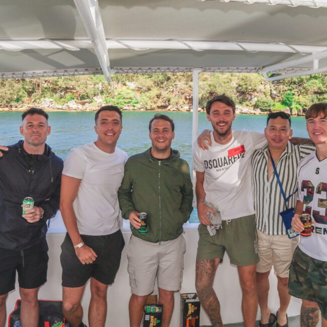 Six men stand smiling on a boat with drinks in hand, posing for a photo. Trees and water are visible in the background through the open side of the boat. It is a sunny day and they appear relaxed and happy.