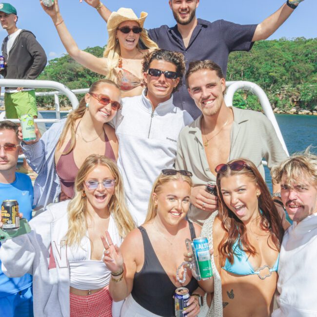 A group of young adults in summer outfits smile and pose on a boat, holding drinks. Green hills and blue sky are in the background, suggesting a festive outdoor gathering near the water.