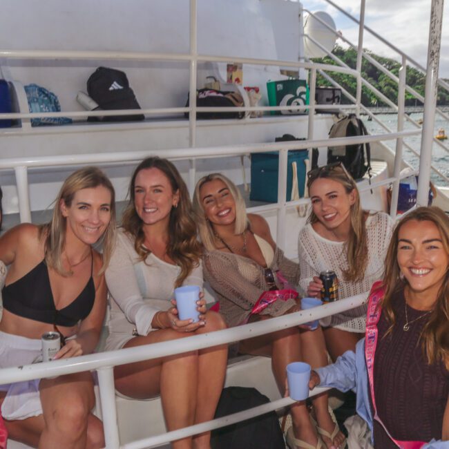 Six women sit and smile together on a boat, holding drinks. They are dressed in casual summer clothes, and the ocean is visible in the background. The mood is relaxed and cheerful.