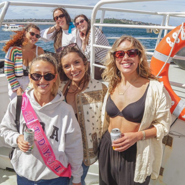 A group of smiling women pose together on a boat, with water and distant buildings in the background. One woman wears a pink "Birthday Girl" sash. Several are holding drinks and wearing sunglasses, enjoying a sunny day.