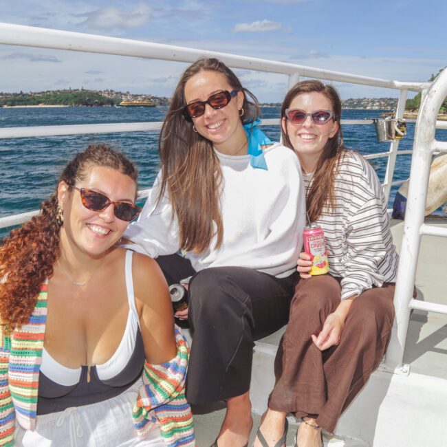 Three women sit and smile on a boat deck under a sunny sky, with blue water and a distant shoreline in the background. One holds a can of drink, and they are dressed in casual, summery clothing.