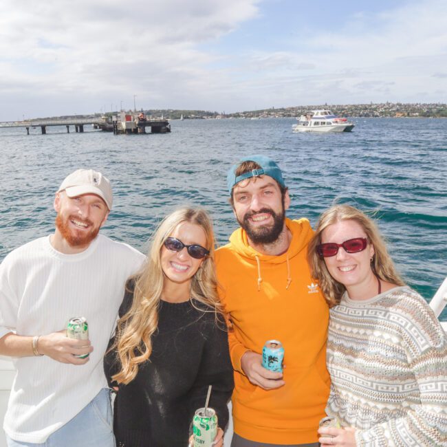 Four friends smile and pose together on a boat, holding drinks. The water, a pier, and a boat are visible in the background under a partly cloudy sky.
