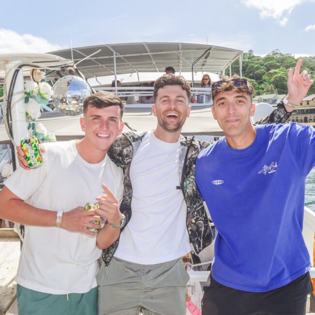 Three young men smile and pose together on a sunny boat party, with water, trees, and buildings in the background; other people are socializing and relaxing on the boat around them.