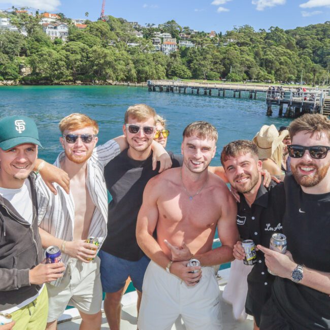 A group of seven smiling men, some shirtless and others in casual clothes, stand close together on a sunny dock by the water. A pier, green trees, and colorful houses are visible in the background.