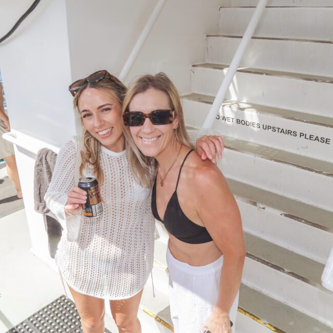 Two women smile at the camera on a boat; one holds a drink and wears a white cover-up over a swimsuit, and the other wears sunglasses and a black bikini top. Stairs and an ocean view are in the background.