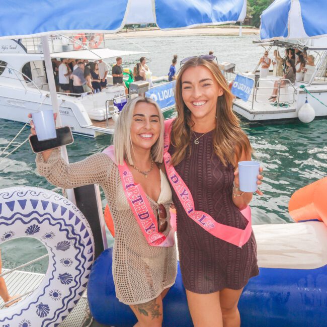 Two smiling women wearing sashes and holding drinks stand on a dock with inflatable floats, with yachts and people in the background on a sunny day.