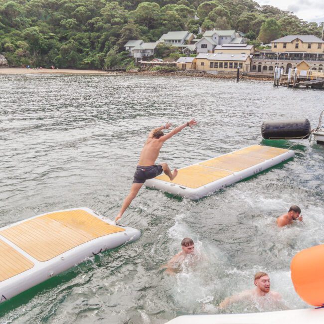 A person leaps off an inflatable dock into the water, joining several others swimming nearby. Boats and buildings are visible in the background by a tree-covered shoreline.