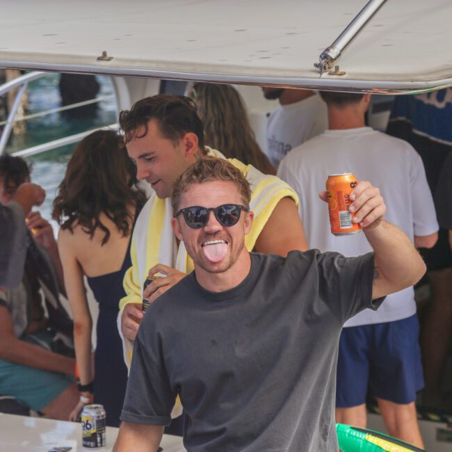 A man in sunglasses and a black shirt stands on a boat, smiling and sticking out his tongue while holding an orange can. Several people are socializing in the background.
