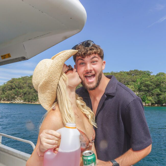 A woman in a sun hat kisses a smiling man on the cheek on a boat. She holds a large drink container, and he holds a can. Blue water and green trees are in the background under a clear sky.