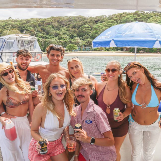 A group of young adults in swimwear smile and pose with drinks on a boat, with a beach, water, and trees in the background. A docked boat and a blue umbrella are also visible. It appears to be a sunny day.