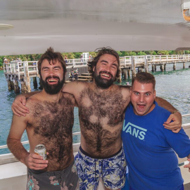 Three men smiling and posing together on a boat; two are shirtless with hairy chests and wet swim trunks, while the third wears a blue VANS shirt. A dock and another boat are visible on the water behind them.