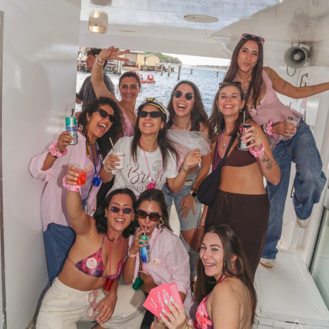 A group of smiling women in casual summer outfits pose together on a boat, holding drinks and party favors, with water and docks visible in the background. Some wear sunglasses and one woman in the center wears a "bride" sash.