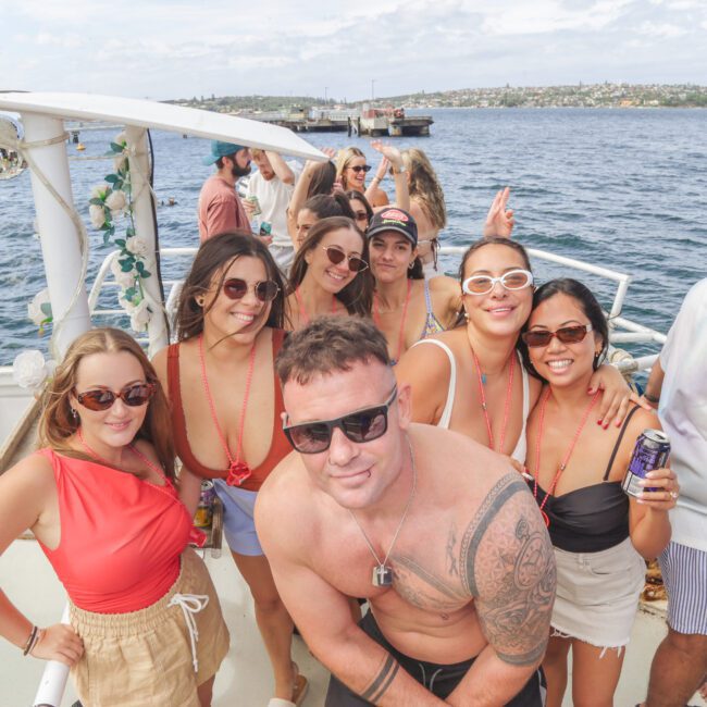 A group of people in summer clothes pose and smile on a boat, with water and a distant shoreline in the background. Some wear sunglasses and hold drinks, enjoying a sunny day outdoors.