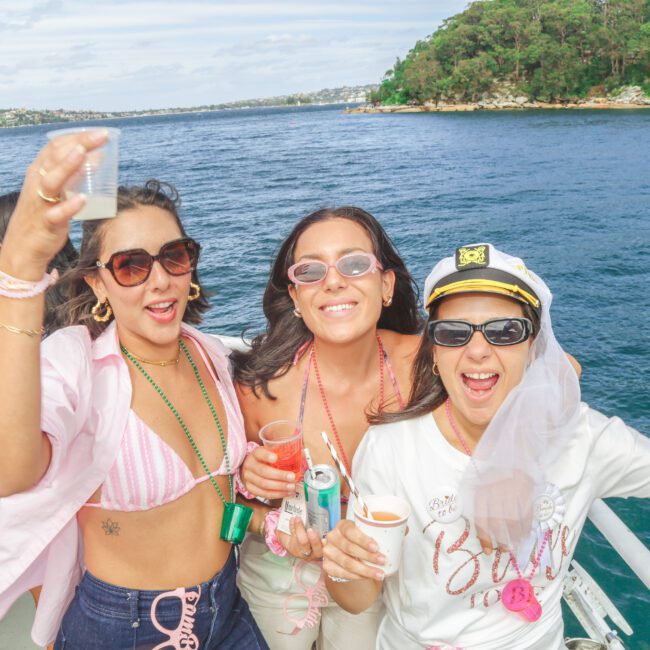 Three women in festive outfits and sunglasses celebrate on a boat, smiling and raising drinks. The woman on the right wears a captain's hat and veil. People and lush green shoreline are visible in the background.