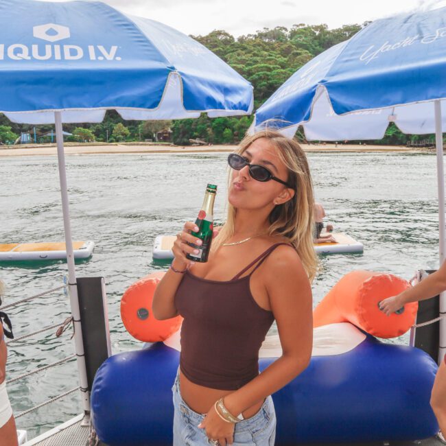 A woman wearing sunglasses, a brown tank top, and jeans stands on a boat holding a bottled drink, making a kissy face. There are blue umbrellas, inflatable seats, and water with trees in the background.