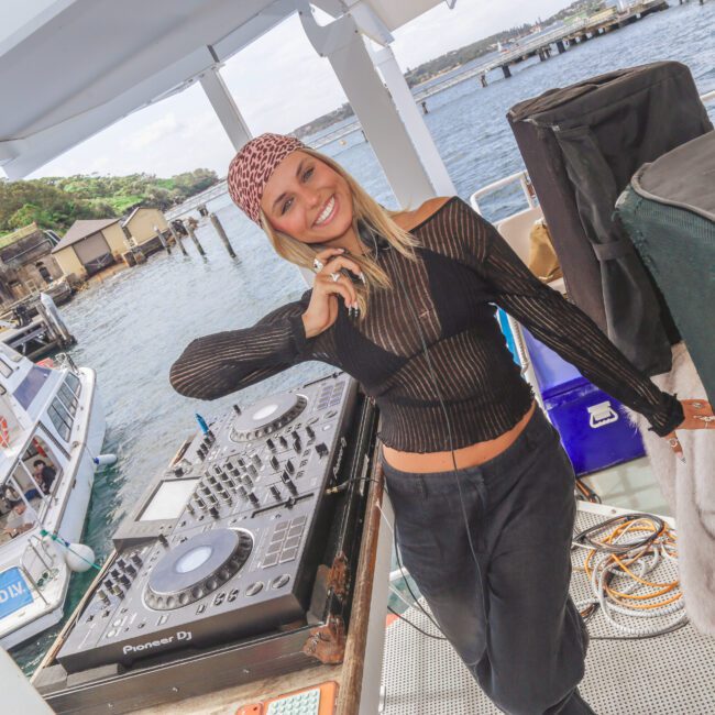 A smiling woman in a black, sheer top and bandana stands next to DJ equipment on a boat, with water, docked boats, and greenery visible in the background.