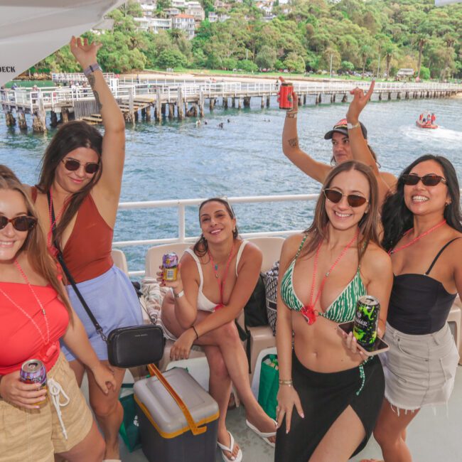 Five smiling women in summer outfits and sunglasses pose with drinks on a boat, with a scenic shoreline, pier, and trees in the background. A "No jumping from upper deck" sign is visible on the wall.