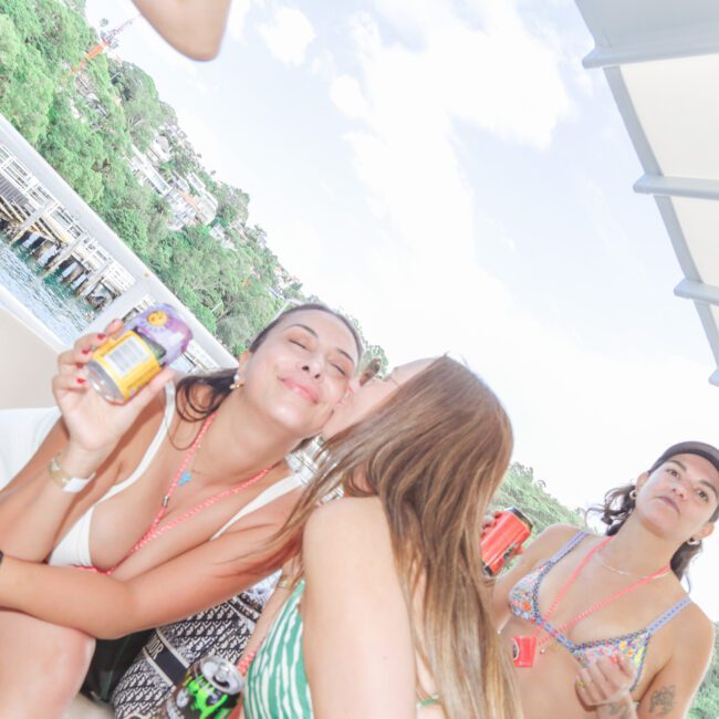 Three women in swimsuits on a boat; one kisses another on the cheek while she smiles holding a drink, and the third woman looks at the camera. Green hills and water are visible in the background.