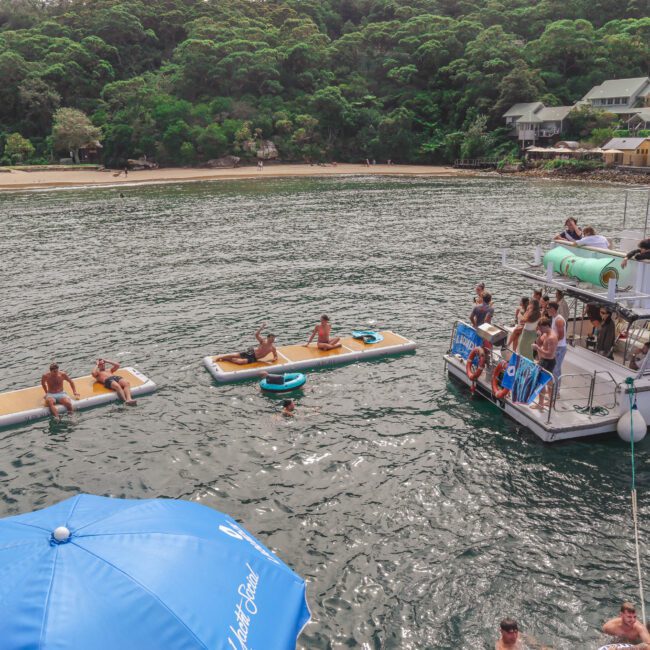 People relax on floating mats and swim near a boat anchored close to a tree-lined shore with buildings. A blue umbrella is visible in the foreground, and the scene has a lively, social atmosphere on the water.