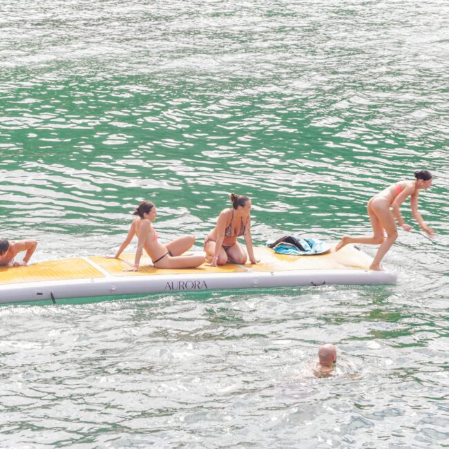 Four people in swimsuits balance on a large floating mat on a green body of water, while others swim nearby. One person is reaching forward, and an inflatable tube is partially visible on the right.