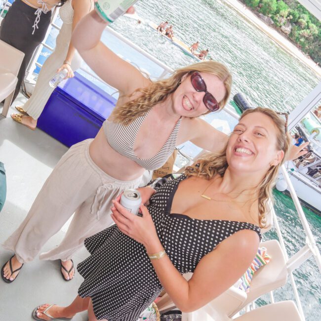 Two women wearing swimsuits and sunglasses smile and hold drinks on a boat, with water and trees in the background. Other people are also visible enjoying the boat trip.