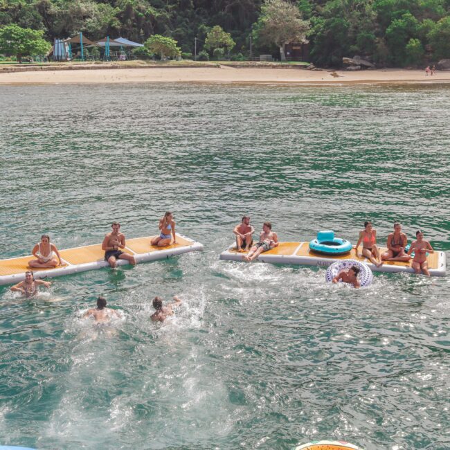 A group of people are swimming and relaxing on floating platforms in green water near a sandy beach with trees in the background. Some are sitting while others play in the water.